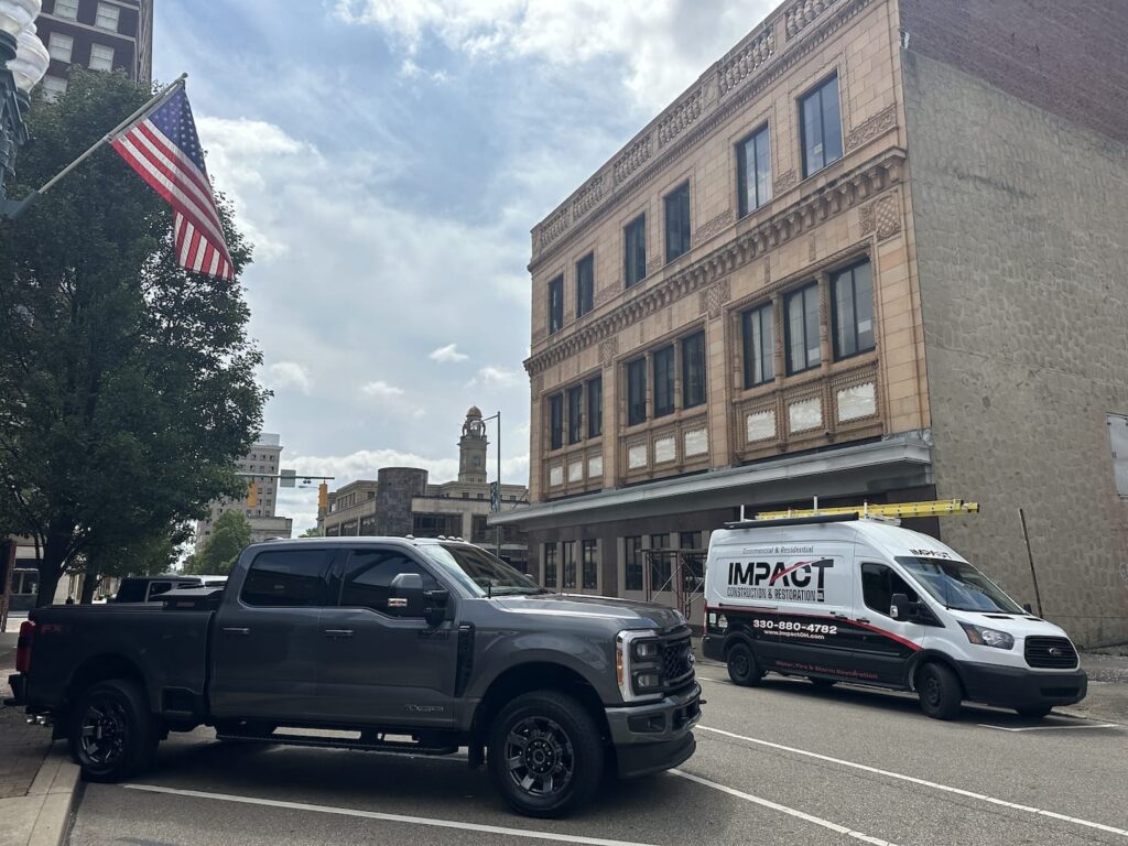 A gray pickup truck and a white van with a Canton Prep School logo are parked on a city street near an old building and an American flag.