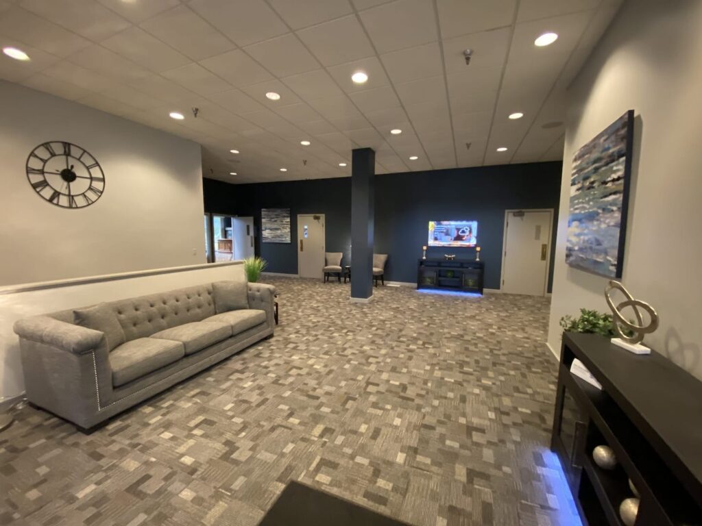A modern office lobby with a gray sofa, a wall-mounted clock, a patterned carpet, and decorative paintings. The lobby also features a black accent wall, two chairs, and a console table with decor.