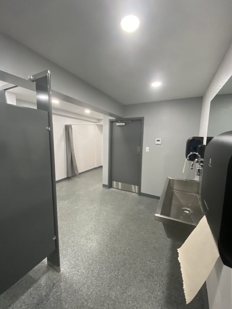 A clean, grey-toned public restroom with a sink, paper towel dispenser, and partitioned toilet stalls.