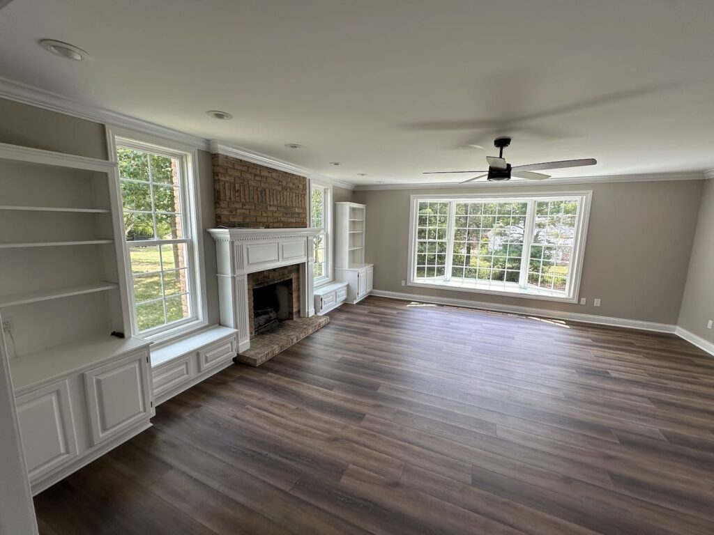 Empty Room With Wood Flooring, A Ceiling Fan, Large Windows, Built In Bookshelves, And A Fireplace With A Brick Surround.