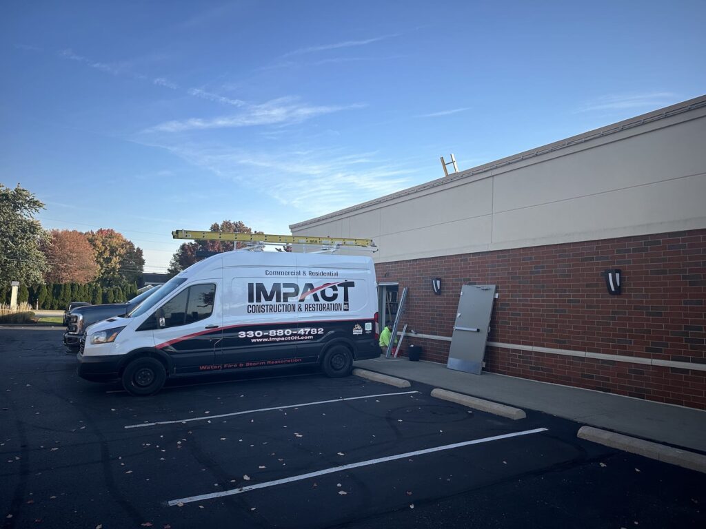 A White Construction Van With "impact Construction & Restoration" Is Parked Beside A Brick Building Under A Clear Blue Sky, Ready For Commercial Door Renovation. Two Ladders Are Attached To The Van's Roof, Showcasing Their Commitment To Excellence.